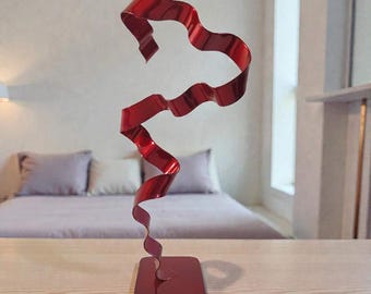 Op de afbeelding: Een moderne, abstracte sculptuur met een vloeiend, lintachtig ontwerp in een levendige rode tint. De sculptuur is gemaakt van metaal en staat op een vierkante basis. Het stuk is ongeveer 46 cm hoog.