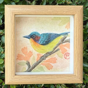 Puede incluir: Una pintura de acuarela enmarcada de un pájaro colorido posado en una rama. El pájaro tiene plumaje azul, amarillo y naranja. La obra de arte está ambientada sobre un fondo claro con acentos florales naranjas y está encerrada en un marco de madera natural.