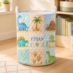 Könnte beinhalten: Ein zylindrischer Aufbewahrungskorb aus Stoff mit gestepptem Muster, das Dinosaurier, einen Vulkan und Palmen zeigt. Der Name ETHAN ist mittig in blauer Schrift aufgedruckt. Der Korb hat zwei weiße Stoffgriffe an der Oberseite.