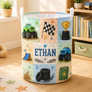 Könnte beinhalten: Ein zylindrischer Aufbewahrungskorb aus Stoff mit weißen Griffen, gesteppter Textur und einem Rastermuster mit Illustrationen eines grünen und eines blauen Monstertrucks, einer Zielflagge, einem goldenen Pokal sowie dem Namen ETHAN.