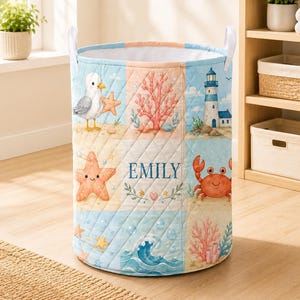 Könnte beinhalten: Ein gesteppter, zylindrischer Wäschekorb mit einem Raster aus maritimen Illustrationen, darunter eine Möwe, Korallen, ein Leuchtturm, ein Seestern, eine Krabbe und Meereswellen. Das mittlere Feld zeigt den Namen EMILY in blauem Text mit einem Herz.
