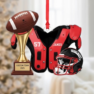May include: A football-themed ornament featuring a brown football, a gold trophy, red and black shoulder pads with the name "JAMES" and number "57", and a black and red helmet. The trophy reads "CUSTOM TEAM 2025".