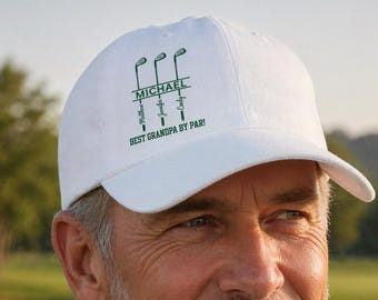 May include: White baseball cap with a curved brim, featuring green golf club graphics and the text "MICHAEL" above "BEST GRANDPA BY PAR!". The cap is worn on a person's head, with a golf course in the background.