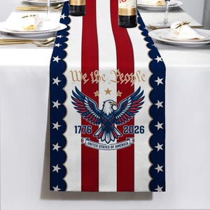 May include: A patriotic table runner with a design featuring red and white stripes, blue borders with white stars, and an eagle emblem. The text "We the People," "1776," and "2026" are also displayed.