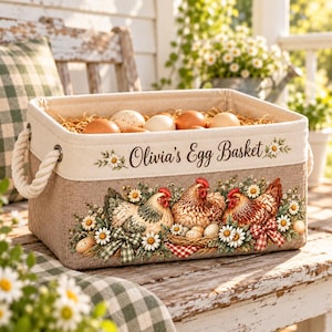 以下が含まれることがあります： ロープハンドル付きの長方形の布製収納バスケット。Olivia's Egg Basketという文字が書かれています。3羽の鶏、デイジー、チェック柄のリボンのイラストが描かれています。バスケットの中には藁と卵がいくつか入っています。