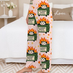 Puede incluir: Pantalones de pijama rosa claro con un patr&oacute;n repetido de zarig&uuml;eyas de dibujos animados en contenedores de basura en llamas. El texto en los contenedores dice "THE HORRORS PERSIST BUT SO DO I". Las llamas naranjas y las estrellas completan el dise&ntilde;o.