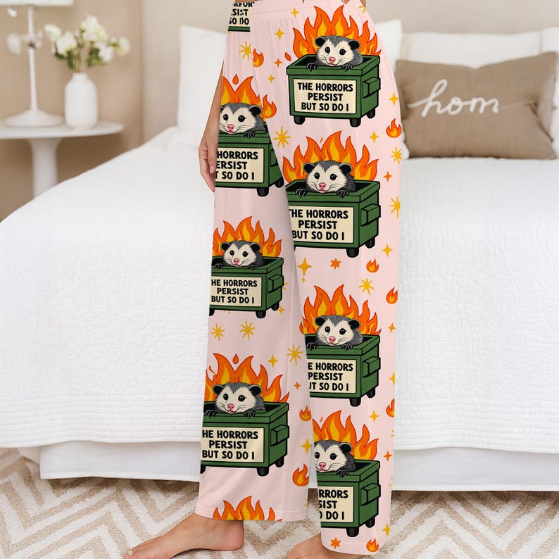 Puede incluir: Pantalones de pijama rosa claro con un patr&oacute;n repetido de zarig&uuml;eyas de dibujos animados en contenedores de basura en llamas. El texto en los contenedores dice "THE HORRORS PERSIST BUT SO DO I". Las llamas naranjas y las estrellas completan el dise&ntilde;o.