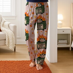 Puede incluir: Pantalones de pijama rosas con un patr&oacute;n repetido de contenedores de basura verdes en llamas, cada uno con una zarig&uuml;eya. El texto en los contenedores dice "THE HORRORS PERSIST BUT SO DO I" y "HORRORS ISIST SO DO I".
