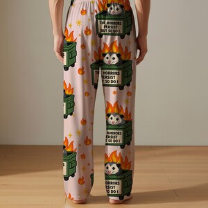 Puede incluir: Pantalones de pijama rosa claro con un patr&oacute;n repetido de contenedores de basura verdes en llamas, zarig&uuml;eyas y estrellas. Cada contenedor de basura tiene el texto "THE HORRORS PERSIST BUT SO DO I". Las zarig&uuml;eyas son grises y blancas.