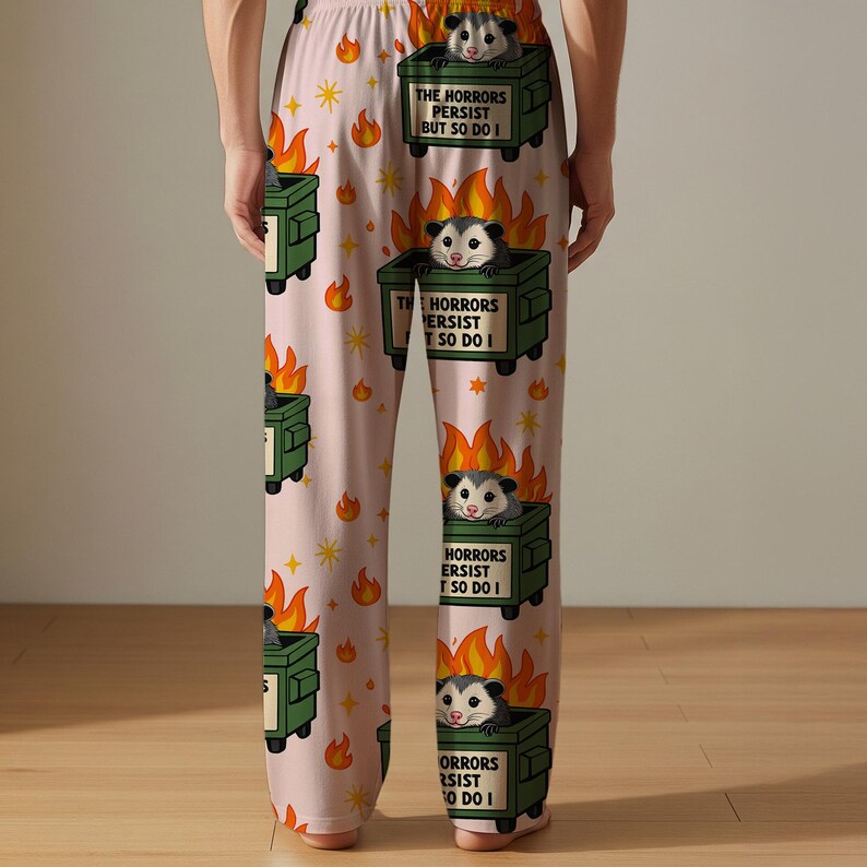 Puede incluir: Pantalones de pijama rosa claro con un patr&oacute;n repetido de contenedores de basura verdes en llamas, zarig&uuml;eyas y estrellas. Cada contenedor de basura tiene el texto "THE HORRORS PERSIST BUT SO DO I". Las zarig&uuml;eyas son grises y blancas.