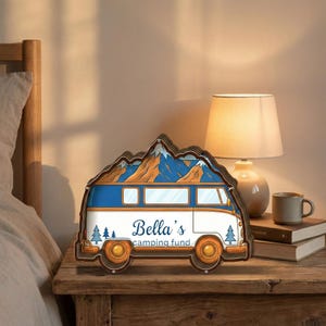 May include: A wooden camper van-shaped money box with a mountain range design on top. The van is white and blue with orange wheels and the text "Bella's camping fund". The money box is on a wooden nightstand.