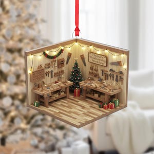 May include: A wooden Christmas ornament shaped like a woodworking shop. The ornament features miniature tools, workbenches, a Christmas tree, and the text "Thomas's Woodworking Workshop." It is decorated with string lights and a red ribbon for hanging.
