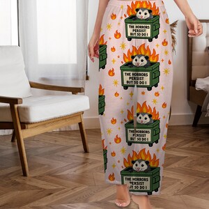 Puede incluir: Pantalones de pijama de color rosa claro con un estampado repetido de contenedores de basura verdes en llamas, cada uno con una zarig&uuml;eya. El texto "THE HORRORS PERSIST BUT SO DO I" est&aacute; impreso en cada contenedor. Estrellas y llamas amarillas completan el dise&ntilde;o.