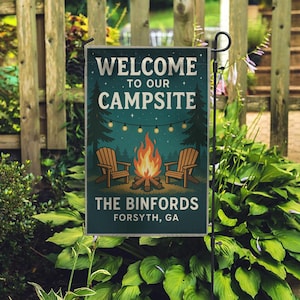 May include: A rectangular garden flag with the text "WELCOME TO OUR CAMPSITE" above an illustration of a campfire, two chairs, and trees. The text "THE BINFORDS FORSYTH, GA" is at the bottom. The flag is displayed outdoors.