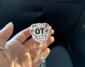 Może przedstawiać: Biała odznaka w kształcie serca z literami "OT" pośrodku, otoczona kolorowymi ikonami i tekstem związanym z terapią zajęciową. Odznaka ma przezroczysty plastikowy klips i chowany sznurek.