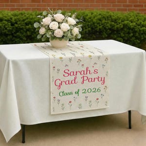 May include: A table set for a graduation party, featuring a white tablecloth and a floral runner. The runner has the text "Sarah's Grad Party Class of 2026" in pink and green. A vase of white roses sits on the table.