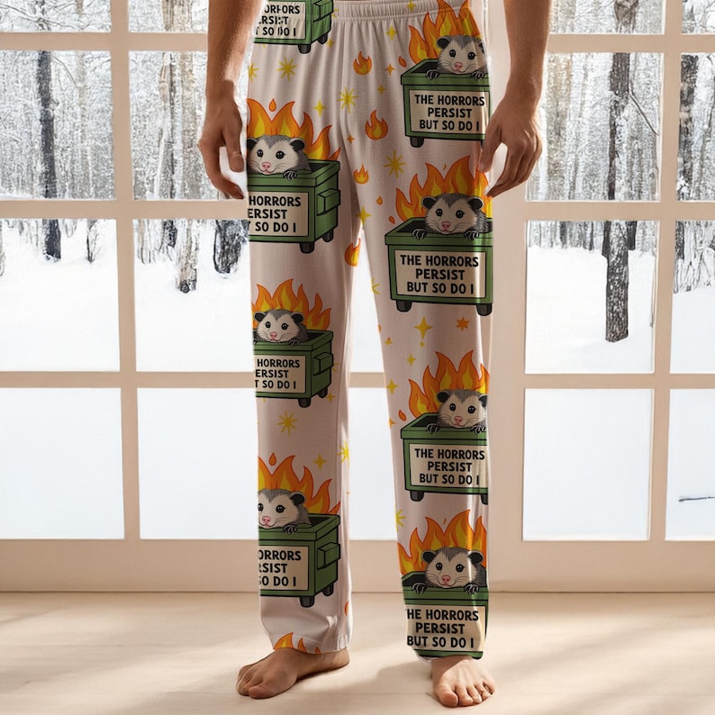 Puede incluir: Pantalones de pijama blancos con un patr&oacute;n repetido de una zarig&uuml;eya en un contenedor de basura en llamas. El texto en el contenedor dice "THE HORRORS PERSIST BUT SO DO I" y "HORRORS PERSIST SO DO I". El dise&ntilde;o incluye estrellas y llamas amarillas.