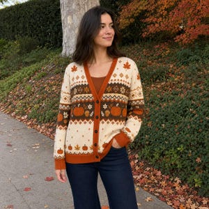 May include: A cream-colored V-neck cardigan featuring a repeating autumn-themed pattern of orange pumpkins, brown acorns, and maple leaves. The garment has a dark brown band across the chest, a solid orange button placket, and orange ribbed cuffs.