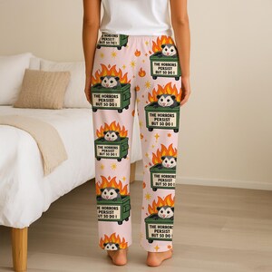Puede incluir: Pantalones de pijama rosas con un patr&oacute;n repetido de una zarig&uuml;eya en un contenedor de basura en llamas. El texto en el contenedor dice "THE HORRORS PERSIST BUT SO DO I". El dise&ntilde;o incluye estrellas y llamas.
