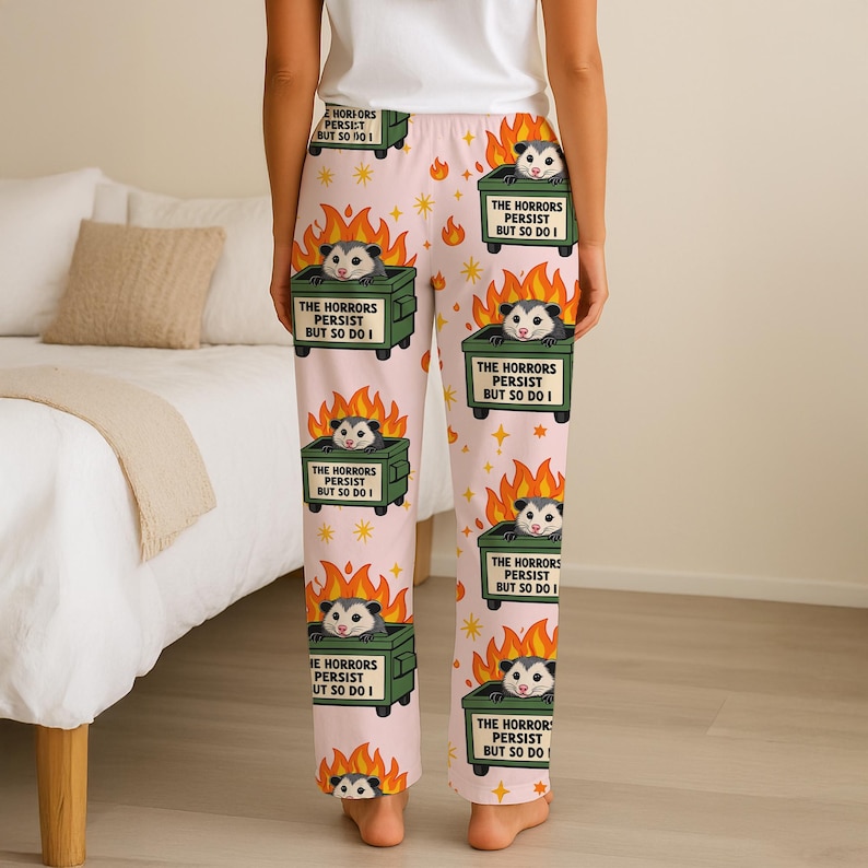 Puede incluir: Pantalones de pijama rosas con un patr&oacute;n repetido de una zarig&uuml;eya en un contenedor de basura en llamas. El texto en el contenedor dice "THE HORRORS PERSIST BUT SO DO I". El dise&ntilde;o incluye estrellas y llamas.
