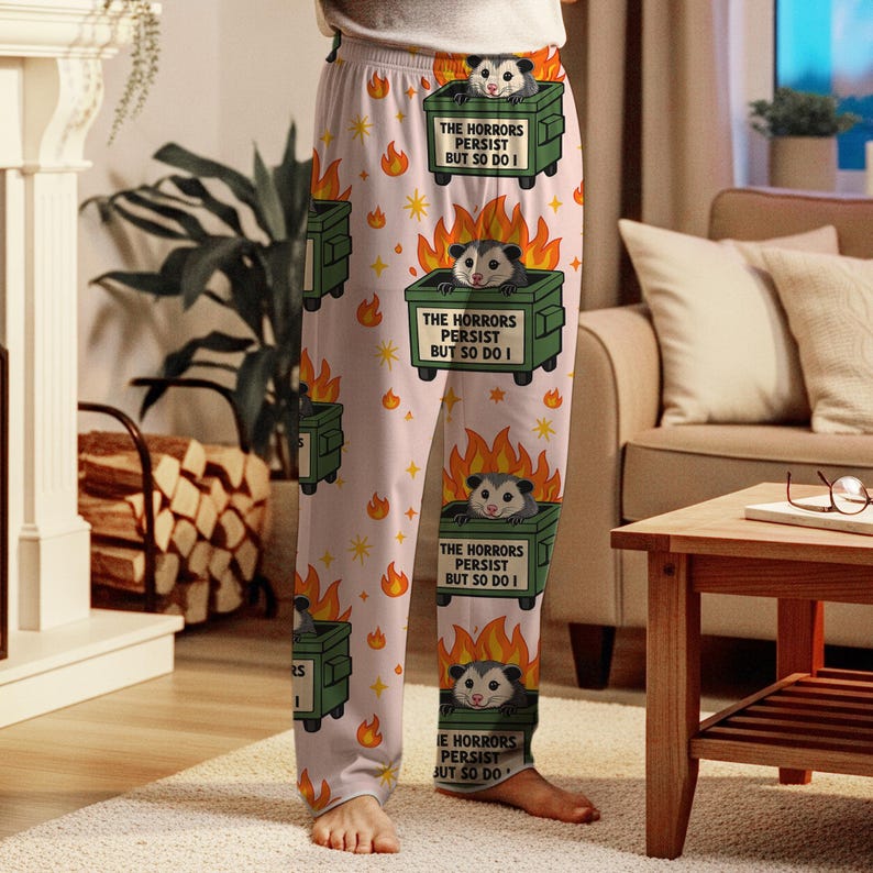 Puede incluir: Pantalones de pijama rosa claro con un estampado repetido de contenedores verdes, zarig&uuml;eyas, llamas y estrellas. Cada contenedor tiene el texto "THE HORRORS PERSIST BUT SO DO I". Los pantalones de pijama son de material suave.