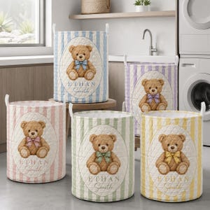 Puede incluir: Cinco cestas de lavandería cilíndricas de tela acolchada con rayas verticales en azul, morado, rosa, verde y amarillo. Cada cesta muestra un parche ovalado central con un oso de peluche marrón con un lazo y el texto Ethan Smith.