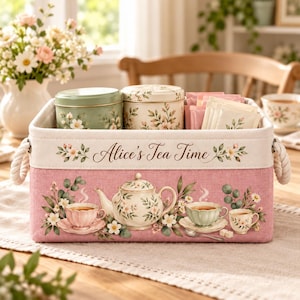 Puede incluir: Una cesta de almacenamiento rectangular de tela rosa con asas de cuerda, con el texto Alice's Tea Time y un estampado floral con tetera y tazas. Dentro hay dos latas metálicas redondas y varios paquetes de té con motivos florales.