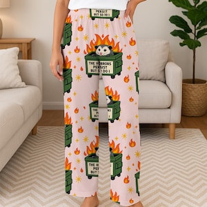 Puede incluir: Pantalones de pijama de color rosa claro con un patr&oacute;n repetido de contenedores verdes con zarig&uuml;eyas, llamas y el texto "THE HORRORS PERSIST BUT S DO I". Los pantalones tambi&eacute;n presentan estrellas y el texto "PERSIST BUT SO I".