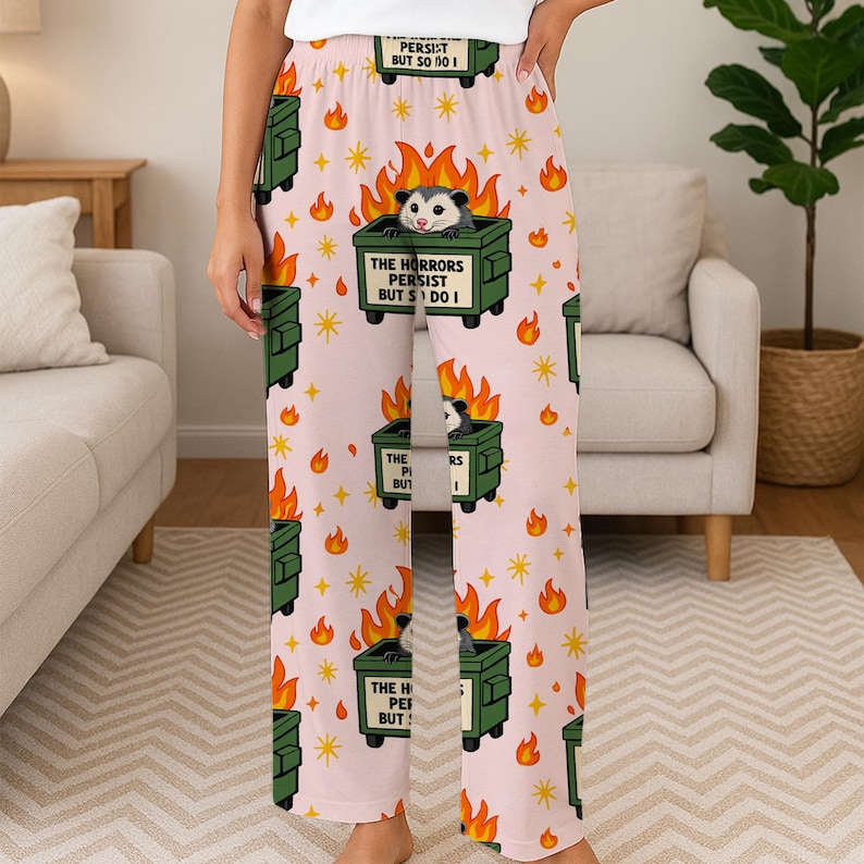Puede incluir: Pantalones de pijama de color rosa claro con un patr&oacute;n repetido de contenedores verdes con zarig&uuml;eyas, llamas y el texto "THE HORRORS PERSIST BUT S DO I". Los pantalones tambi&eacute;n presentan estrellas y el texto "PERSIST BUT SO I".