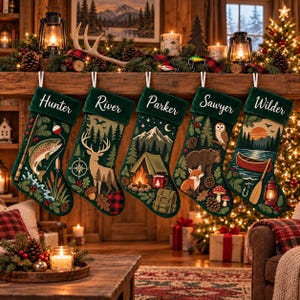 Op de afbeelding: Vijf donkergroene kerstsokken hangen op een rij, elk gepersonaliseerd met witte schrijfletter-namen: Hunter, River, Parker, Sawyer en Wilder. De sokken bevatten illustraties met een outdoor-thema, waaronder een vis, hert, kampvuur, beer en kano.