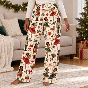 Pode incluir: Calças de pijama cor de creme com um padrão repetitivo de quebra-nozes, bailarinas em vestidos vermelhos, verdes e azuis, tambores, bengalas doces e presentes. O nome AMARA está impresso em pequenas faixas por todo o design festivo.