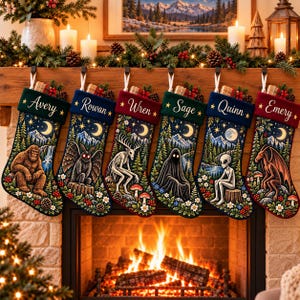 Op de afbeelding: Zes kerstsokken hangen op een rij, elk met een unieke illustratie van een cryptide zoals Bigfoot, Mothman, een wendigo, een geest, een alien en een draak, met de namen Avery, Rowan, Wren, Sage, Quinn en Emery geborduurd op de boorden.