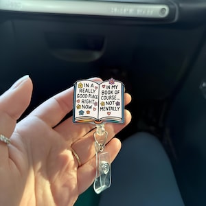May include: A badge reel with a book design. The book has the text "In a really good place... right now" on the left page and "In my book of course... not mentally" on the right page. The badge reel is white, with a clear plastic clip.