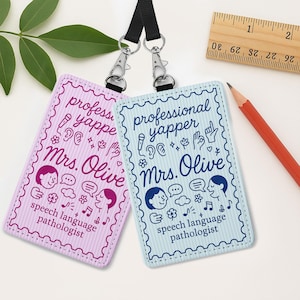 Puede incluir: Dos tarjetas de identificación rectangulares con cordones negros. Una es rosa con el texto "professional yapper Mrs. Olive speech language pathologist". La otra es azul claro con el mismo texto. Una regla y un lápiz están en el fondo.