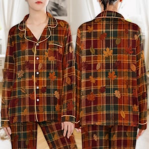 Puede incluir: Una camisa de pijama de manga larga con botones, con un patrón de cuadros en tonos rojo oscuro, naranja y verde, acentuado con un estampado de hojas de otoño y ribetes amarillos finos en el cuello, los puños y la tapeta frontal.