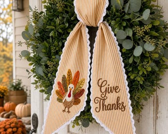May include: A decorative fabric door hanger featuring two long, tan and white striped tails with white lace trim. One tail displays an embroidered turkey with colorful feathers, while the other tail features the embroidered text Give Thanks.