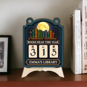 Może przedstawiać: Drewniany znak na biurko z ciemnoniebieską obwódką i jasną drewnianą podstawą. Na znaku widnieje napis "BOOKS READ THIS YEAR" nad liczbami "315". Tło przedstawia pełnię księżyca i rząd książek. Tekst "EMMA'S LIBRARY" znajduje się na dole.