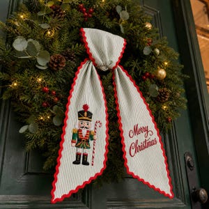 May include: A decorative fabric bow with a light grey and white pinstripe pattern and red scalloped edges. One tail features an embroidered nutcracker holding a candy cane, while the other tail displays the red embroidered text Merry Christmas.