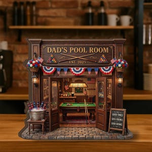 Może przedstawiać: Miniaturowy drewniany model "Dad's Pool Room" z wnętrzem. Fasada posiada szyld, amerykańskie flagi i dekoracyjne girlandy. Wewnątrz widoczny jest stół bilardowy, bar i inne detale. Znak głosi "Good Times, Good Friends, Good Drinks".
