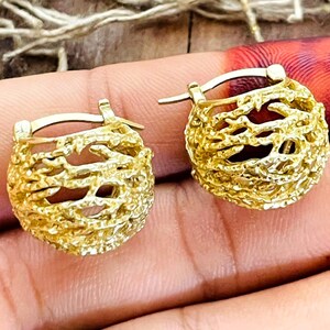 May include: A pair of gold-toned hoop earrings featuring a textured, organic, branch-like design. The earrings have a rounded, cage-like structure with a polished finish and a hinged closure mechanism, showcasing a detailed, intricate, and porous surface.