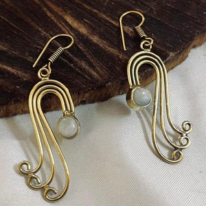 May include: A pair of gold-colored earrings with a unique design. Each earring features a curved, layered structure with a small, round, light-colored stone at the center and decorative swirls at the bottom. The earrings are displayed on a dark wooden surface.