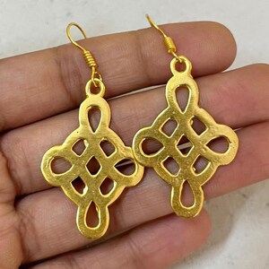 May include: A pair of gold-colored earrings with an openwork design. Each earring features a symmetrical, geometric pattern with a central knot-like shape and curved edges. The earrings are displayed against a neutral background.