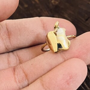 Puede incluir: Un anillo dorado con un colgante en forma de diente estilizado. El diente tiene un relleno de esmalte blanco y un pequeño lazo en la parte superior. El anillo se sostiene en una mano sobre un fondo de madera oscura.