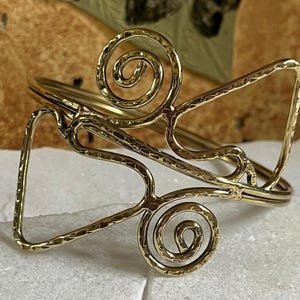 Può includere: Un bracciale a polsino in metallo color oro con un design aperto. Il bracciale presenta una texture martellata e volute decorative. Il bracciale è appoggiato su una superficie bianca.