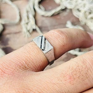 May include: A silver-colored signet ring with a rectangular face and a black vertical line design. The ring is worn on a finger, showcasing its geometric shape and minimalist style. The ring is a piece of jewelry.
