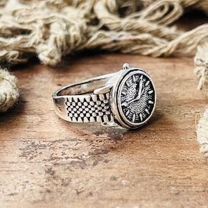 Puede incluir: Anillo plateado con diseño de esfera de reloj. El anillo presenta una esfera de reloj detallada con marcadores de hora y manecillas, y una banda diseñada para parecerse a una correa de reloj. El anillo está sobre una superficie de madera rústica.