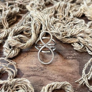 May include: A silver-colored ring with an infinity symbol design. The ring has a layered appearance with multiple bands. The background features a wooden surface and strands of beige twine.
