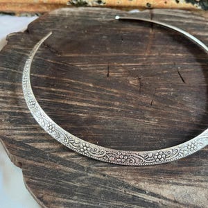 Könnte beinhalten: Ein silberfarbenes Choker-Halsband mit einem aufwendigen Blumenmuster. Die Halskette ist ein starres, offenes Kragendesign, das auf einer dunklen Holzoberfläche liegt. Das detaillierte Design weist kleine, sich wiederholende Blumen- und Punktmotive auf.