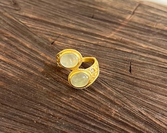 Può includere: Un anello color oro con due pietre preziose ovali verde chiaro. L'anello ha un design ornato con dettagli decorativi attorno alle pietre preziose e lungo la fascia. L'anello è esposto su una superficie di legno scuro.