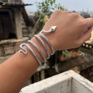 Op de afbeelding: Een zilverkleurige slangenarmband die om een pols gewikkeld is. De armband heeft een gedetailleerde slangenkop en -staart, waarbij het lichaam van de slang zich om de arm wikkelt. De armband is gemaakt van een getextureerd metaal.